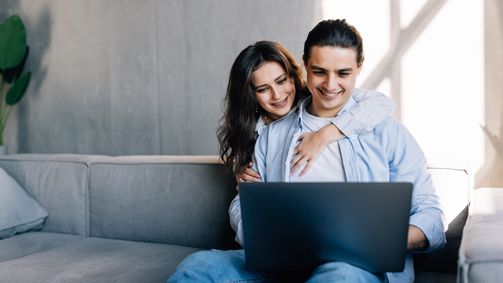 Young couple surfing on internet with laptop sitting on couch CPE e1734730830477